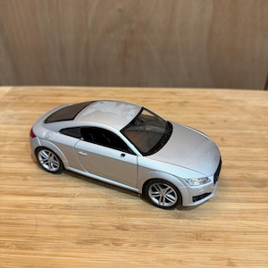May include: Silver Audi TT model car with black windows and wheels. The toy car is on a wooden surface. The car has the Audi logo on the front.