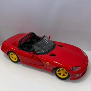 Puede incluir: Un coche descapotable de juguete rojo con interior negro y ruedas amarillas. El coche tiene un diseño elegante y un parabrisas. El coche es una réplica en miniatura de un coche deportivo.