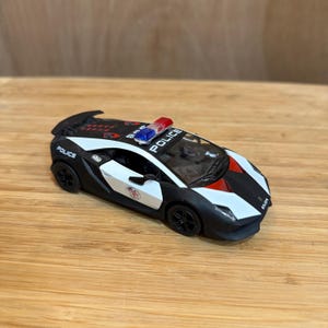 May include: A miniature police car in black, white, and red, with the word "POLICE" visible. It features a red and blue light bar and black wheels. The toy car is set on a wooden surface.