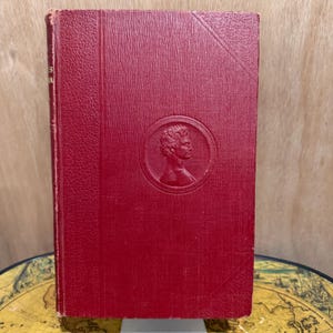 May include: A vintage, red hardcover book with a textured cover. The book features a circular embossed portrait on the front cover. The spine shows the title in faded gold lettering. The book is displayed on a wooden surface.