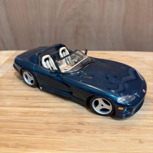 Puede incluir: Un coche de juguete descapotable de color verde azulado oscuro con asientos blancos y ruedas plateadas. El coche tiene un diseño elegante con un perfil bajo y un parabrisas. El coche de juguete está sobre una superficie de madera.