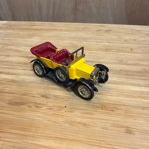 Può includere: Un'auto giocattolo vintage gialla con interni rossi. L'auto ha pneumatici neri con cerchi dorati e un telaio nero. L'auto ha una griglia anteriore dorata e un telaio del parabrezza.
