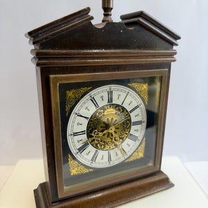 May include: A vintage brown wooden clock with a decorative top finial. The clock face is white with black Roman numerals and gold filigree accents. The clock has a rectangular shape and a glass front.