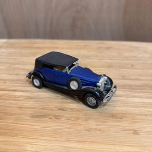 May include: A miniature blue and black vintage car with a black convertible top. The toy car has silver accents on the front and wheels. The car is sitting on a wooden surface.