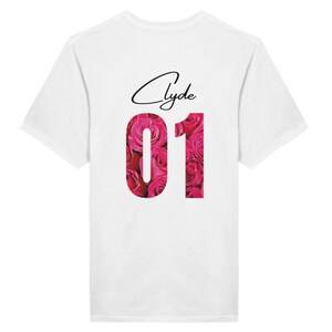 Könnte beinhalten: Weißes T-Shirt mit einem rosafarbenen Blumendesign und der Zahl "01" in Weiß. Der Name "Clyde" ist in schwarzer Kursivschrift über der Zahl geschrieben.