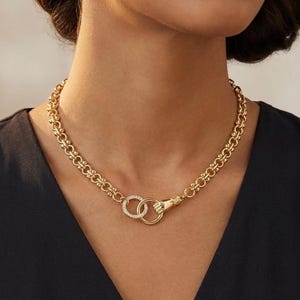 May include: A gold-toned necklace with a unique design. The necklace features a chain-link style with a hand clasp and two interlocking rings, one of which is adorned with small, clear stones. The necklace is worn against a dark background.