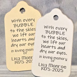 May include: Two memorial tags with a scalloped top and a hole for hanging. The tags are cream and white, with black text that reads "With every bubble to the skies, we lift our hearts and dry our eyes. In loving memory of Lisa Monroe 1975-2025."