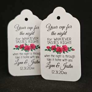 May include: Two white favor tags with a floral design featuring red roses. The text on the tags reads "Your cup for the night for WHATEVER TASTES RIGHT!" and "when the night is through take it home with you Lynn & Justin 12.31.20xx".