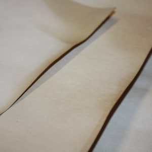 May include: Close-up of two sheets of light tan paper with darker edges. The paper has a smooth texture and is slightly curved, suggesting flexibility. Soft shadows from the lighting highlight the paper's subtle variations. The paper is likely A4 size.