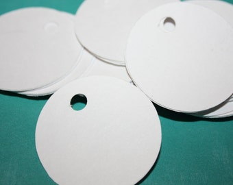 May include: A stack of white round paper tags with a hole punched in the center. The tags are on a teal surface.