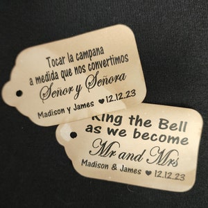 May include: Two brown wooden tags with black text. The top tag reads "Tocar la campana a medida que nos convertimos Señor y Señora Madison y James 12.12.23". The bottom tag reads "Ring the Bell as we become Mr and Mrs Madison & James 12.12.23".