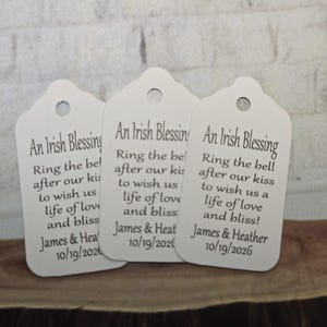 May include: Three white gift tags with scalloped edges. Each tag has a hole at the top and features the text "An Irish Blessing" followed by a poem and names with a date. The tags are on a wooden surface.