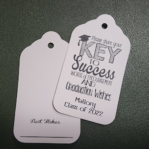 May include: White gift tag with black text that reads "Please share your KEY to Success Words of encouragement and Graduation Wishes Mallory Class of 2022". The tag has a hole punched in the top and a black line for writing.