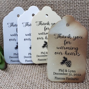 May include: Four rustic wedding favor tags with a pine cone design and the text "Thank you for warming our hearts". The tags are made of brown paper and have a distressed look. The bottom tag includes the names "Phil & Lynn" and the date "December 31, 2022".