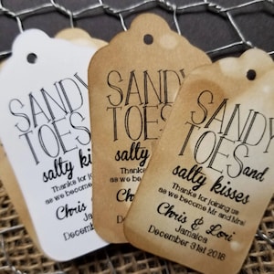 May include: Three brown paper favor tags with the text "Sandy Toes Salty Kisses" and the date "December 31st 2018". The tags are tied with twine and have a distressed look.