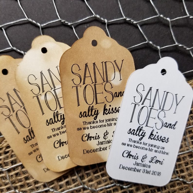 May include: Three brown paper favor tags with the text "Sandy Toes and Salty Kisses" and the wedding date of December 31st, 2018. The tags are tied with twine and have a hole punched at the top.