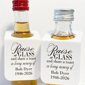 May include: Two miniature liquor bottles with red and silver caps, each in a white holder. The holders have the text "Raise a GLASS and share a toast in loving memory of Bob Dyer 1946-2026" in black script.