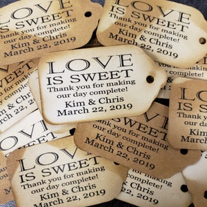 May include: A stack of brown paper favor tags with the text "Love is Sweet" printed in black. The tags are decorated with a black border and include the text "Thank you for making our day complete! Kim & Chris March 22, 2019".