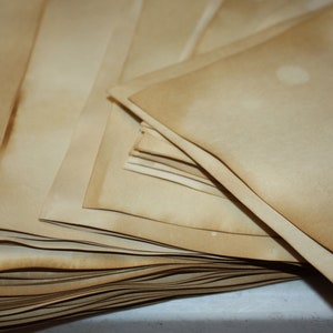 May include: A pile of aged, tan-coloured paper sheets. The edges of the paper have a darker, browned appearance, suggesting an antique or vintage aesthetic. The paper is stacked in a neat pile.