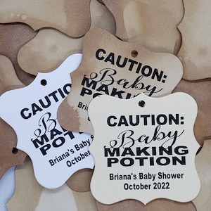 May include: A set of four brown and white paper favor tags with the text "Caution: Baby Making Potion" in black. The tags are shaped like a heart and have a hole at the top for hanging. The text also includes the name "Briana's Baby Shower" and the date "October 2022".