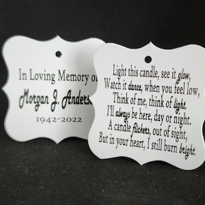 May include: Two white memorial candle tags with black text. The left tag reads "In Loving Memory of Morgan J. Anders 1942-2022". The right tag reads "Light this candle, see it glow, Watch it dance, when you feel low, Think of me, think of light. I'll always be here, day or night. A candle flickers, out of sight, But in your heart, I still burn bright."