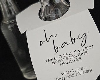 May include: White baby onesie shaped bottle tag with black text that reads "oh baby take a shot when baby stevens arrives with love, amy and michael"