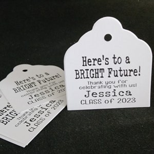 May include: White graduation favor tags with black text. The tags read "Here's to a BRIGHT Future!" and "Thank you for celebrating with us! Jessica CLASS of 2023".