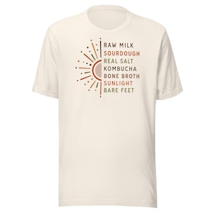 May include: Cream-colored t-shirt featuring a sunburst design in shades of brown and orange. The text includes "RAW MILK, SOURDOUGH, REAL SALT, KOMBUCHA, BONE BROTH, SUNLIGHT, BARE FEET". A casual, comfortable tee.
