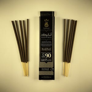 Pode incluir: Uma embalagem preta de incenso "SCENTS OF MECCA" com o texto "INCENSES STICKS". A embalagem tem detalhes dourados e contém 90 varetas. Feixes de varetas de incenso são dispostos em ambos os lados da embalagem.