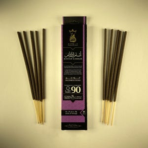 Puede incluir: Palitos de incienso de color marrón oscuro con tallos claros, dispuestos sobre un fondo claro. Una caja morada y negra en el centro muestra texto árabe, las palabras "SCENTS OF ALANDALOS" e "INCENSES STICKS". La caja indica un tiempo de combustión de 90 minutos.