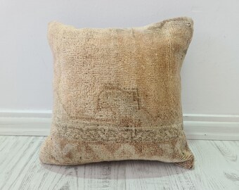 May include: Square beige pillow with a textured, woven appearance. The pillow has a faded, vintage look with subtle patterns in shades of brown and pink. The pillow is placed against a white wall and light-colored wooden floor.