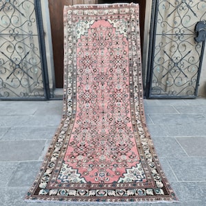 Op de afbeelding: Een rechthoekig, vintage tapijt met een roze middenveld en ingewikkelde geometrische patronen in bruin, crème en blauw. Het tapijt heeft een gedetailleerde rand en wordt getoond op een stenen oppervlak.