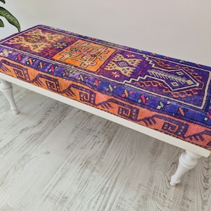Könnte beinhalten: Eine weiße Bank mit gedrehten Beinen hat ein farbenfrohes, gemustertes Kissen. Das Kissen ist ein traditioneller türkischer Teppich in Orange, Lila und Blau.