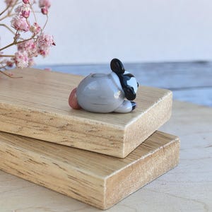 May include: A small glass figurine of a raccoon, lying on its side with its head resting on its paws. The raccoon is gray with black markings around its eyes and a black tail. The figurine is hand-blown and has a unique, whimsical style.
