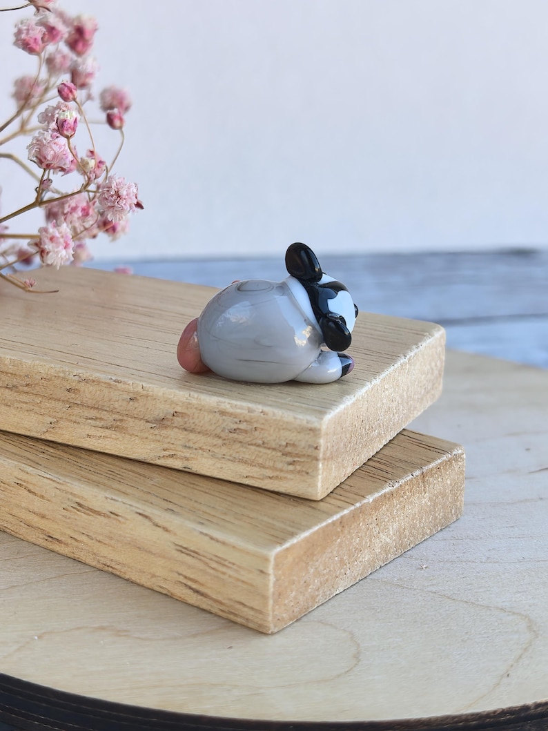 May include: A small glass figurine of a raccoon, lying on its side with its head resting on its paws. The raccoon is gray with black markings around its eyes and a black tail. The figurine is hand-blown and has a unique, whimsical style.