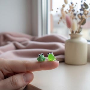 May include: Two small, green frog figurines with black eyes are displayed on a finger. One frog is solid green, the other is translucent. The frogs are miniature and whimsical, likely decorative items or charms.