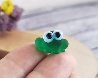 May include: A small green glass frog figurine with large blue eyes. The frog is sitting on a finger.