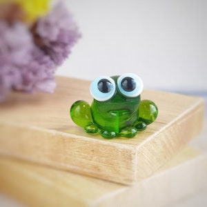 May include: A small green glass frog figurine with large white eyes and black pupils. The frog is sitting on a wooden surface.