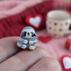 May include: A small, gray sloth figurine with black accents around the eyes and a white belly. The sloth is in a sitting position with its arms and legs tucked in. The figurine is held in a hand.