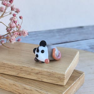 May include: A small glass figurine of a possum, lying on its side with its tail curled up. The possum is white with black eyes and a black nose, and its tail is pink.