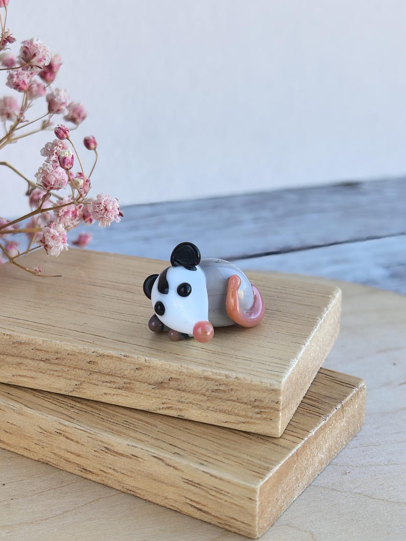 May include: A small glass figurine of a possum, lying on its side with its tail curled up. The possum is white with black eyes and a black nose, and its tail is pink.