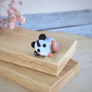 May include: A small glass figurine of a possum, with black and white markings, lying on its side. The figurine is made of clear glass with a white and black swirl pattern. The possum has a pink nose and a pink tail.