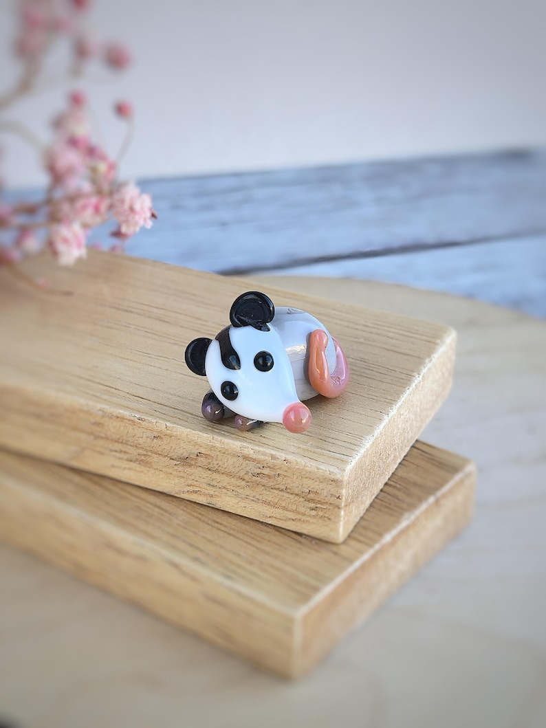 May include: A small glass figurine of a possum, with black and white markings, lying on its side. The figurine is made of clear glass with a white and black swirl pattern. The possum has a pink nose and a pink tail.