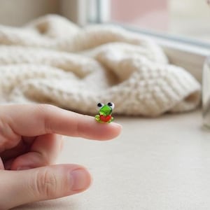May include: A tiny glass frog figurine with a green body, black eyes, and a red heart. The frog is perched on a fingertip, showcasing its small size and intricate details. The background is softly blurred, highlighting the figurine.