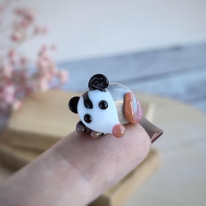 May include: A small glass figurine of a panda bear. The panda is white with black markings and has a pink nose. The figurine is sitting on a finger.