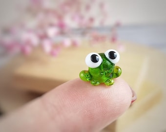 May include: A small green glass frog figurine with large white eyes and black pupils. The frog is sitting on a finger.