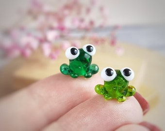 May include: Two small green glass frogs with white eyes. The frogs are sitting on a finger.