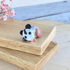 May include: A small glass figurine of a possum with black and white stripes, brown eyes, and a pink nose. The possum is lying on its side with its tail curled up. The figurine is on a wooden surface.
