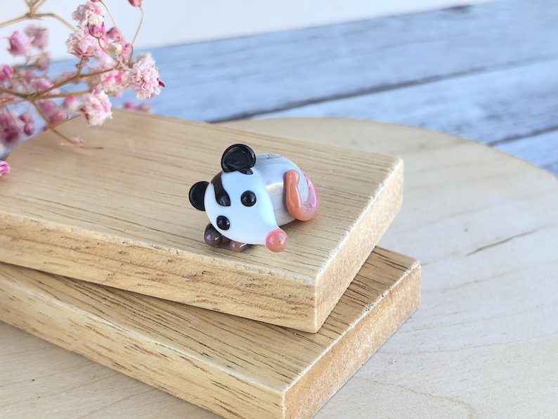 May include: A small glass figurine of a possum with black and white stripes, brown eyes, and a pink nose. The possum is lying on its side with its tail curled up. The figurine is on a wooden surface.