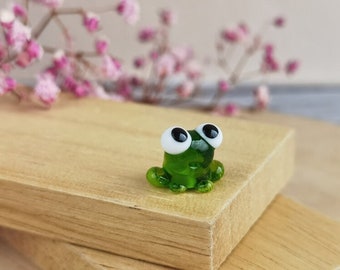 May include: A small green glass frog figurine with white eyes. The frog is sitting on a wooden surface.